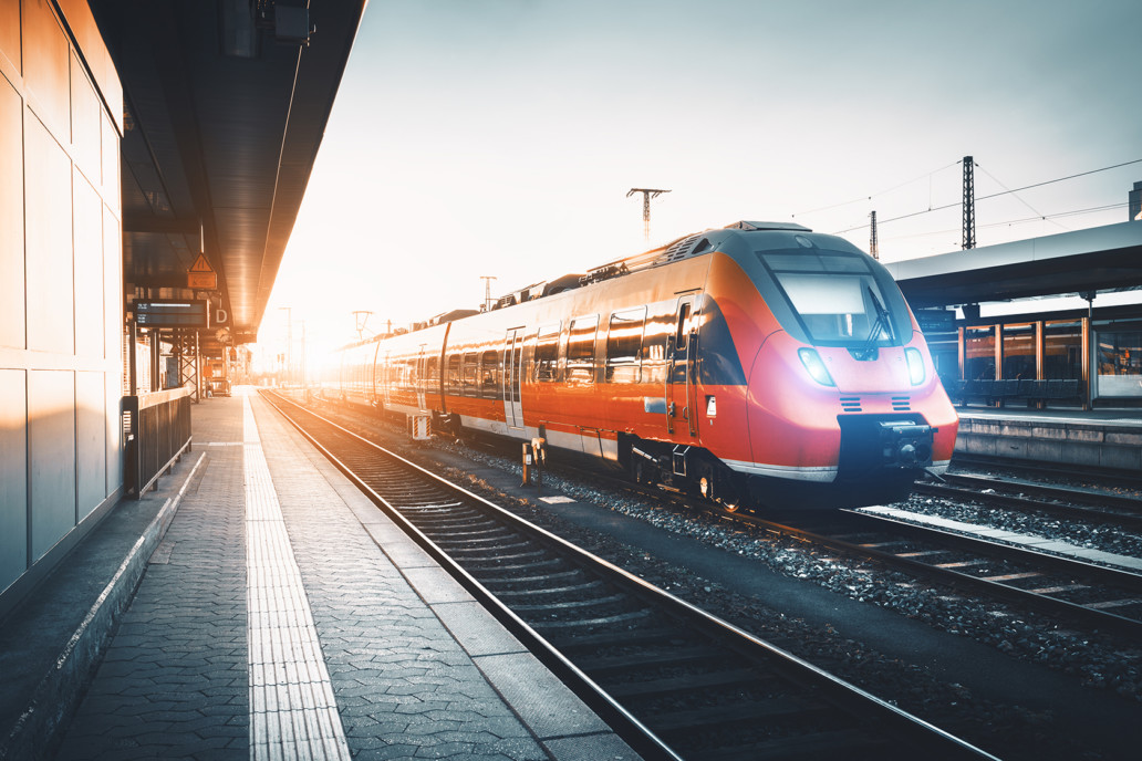 Modern high speed red commuter train at the railway station at sunset. Turning on train headlights. Railroad with vintage toning. Train at railway platform. Industrial landscape.